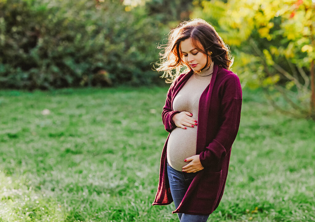 schwangere Frau steht auf einer grünen Wiese draußen und umfasst ihren Bauch
