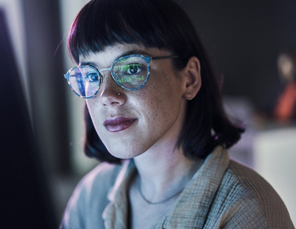 junge Frau sitzt vor einem Bildschirm, Zahlen spiegeln sich in ihrer Brille